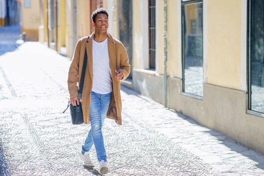 Young black man walking down the street carrying a briefcase and a smartphone. Cuban guy in urban background.