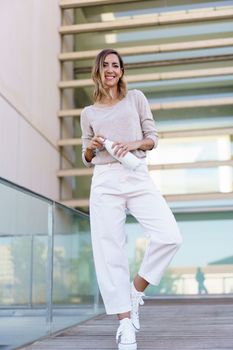 Full body happy female in stylish clothes, opening bottle of water and looking at camera with smile while standing on bridge with glass railing outside contemporary building