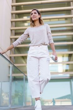 Low angle of stylish female with bottle of water touching glass railing and looking away while walking on bridge outside contemporary building