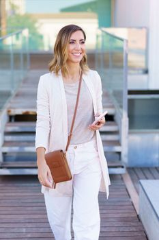 Middle-aged woman walking near an office building carrying a handbag and a smartphone. Caucasian female in urban background.