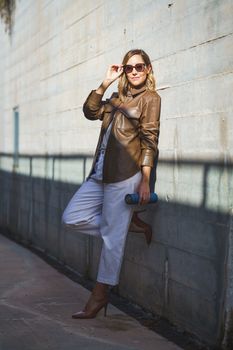 Full body self assured female in stylish clothes with thermos looking at camera and adjusting sunglasses while leaning on sunlit wall on city street