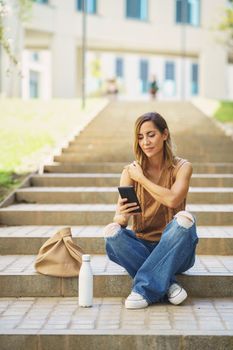 Full body woman in stylish clothes touching shoulder, and reading news on cellphone while sitting on steps near bottle of water and cotton bag on sunny day on street