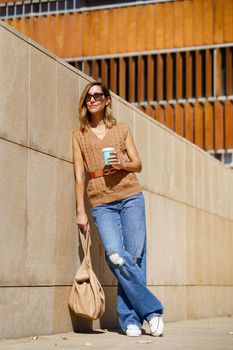 Full body adult woman with stylish bag and takeaway cup of coffee leaning on modern wall and looking away on sunny day on city street