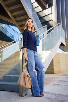 Full body female in stylish clothes with bag and thermos leaning on glass railing of stairs outside modern building in daytime