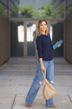 Full body merry woman in stylish clothes with bag and thermos looking at camera with smile while walking outside contemporary building
