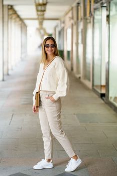 Full body of positive young female in beige blouse and pants with shoulder bag looking at camera smiling