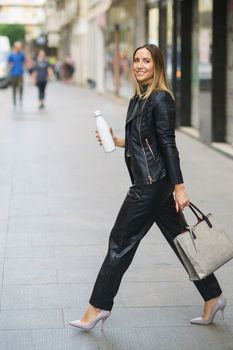 Side view of confident stylish woman in leather outfit and heels carrying modern handbag with eco friendly bottle and walking in city