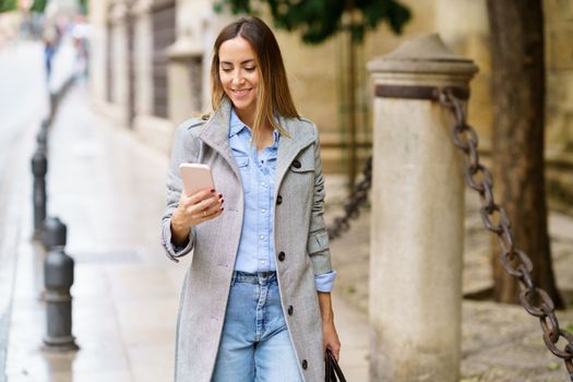 Positive adult female in casual clothes and stylish coat scrolling messages on mobile phone while walking on pavement outside historic building in daytime in city