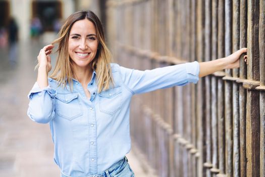 Happy adult female in stylish blue shirt looking at camera with smile and touching hair while grasping metal fence on blurred background of city street