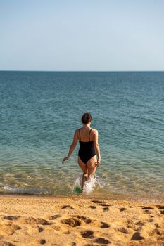 Woman sea swimsuit sand. A girl in full growth stands with her back and enters the sea in a black swimsuit. Alone on the beach on a sunny day