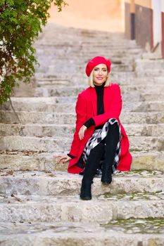 Full length of graceful young female tourist, with blond hair in fashionable red beret and coat sitting on old stone stairway and smiling while resting during sightseeing in town