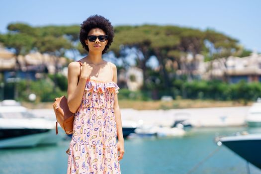 Young black woman in dress and sunglasses with curly hair, carrying bag on shoulder and looking at camera while standing on pier on blurred background of harbor in resort city