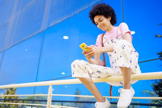 Glad African American female with Afro hairstyle in overall browsing cellphone while sitting on railing near glass building in city