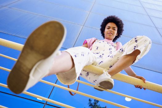 From below full body of young African American female in casual outfit looking at camera with serious face while sitting on railing