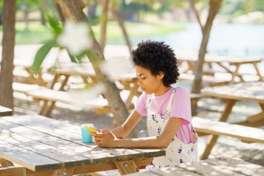 Side view of focused African American female texting message on cellphone while sitting at wooden table in park on summer day
