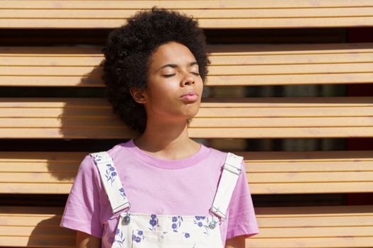 Charming African American female in casual wear standing under bright sunlight with closed eyes near wooden wall on street on summer day