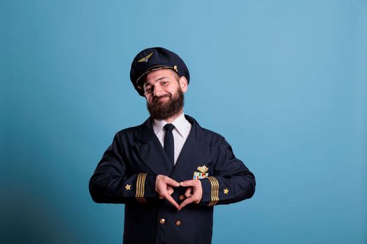 Happy airplane captain showing heart shaped love symbol with fingers, conceptual romance gesture. Friendly airliner aviator in uniform expressing affection feelings front view medium shot