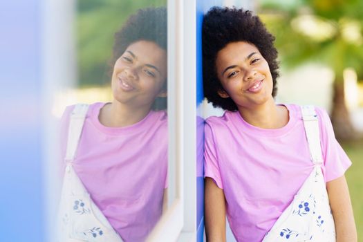 Happy African American female in casual clothes looking at camera while standing near window of house on street against blurred background