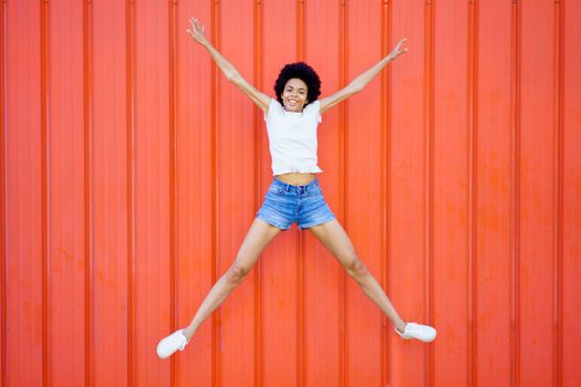 Full body of optimistic African American female in casual clothes looking at camera while star jumping on red background in city