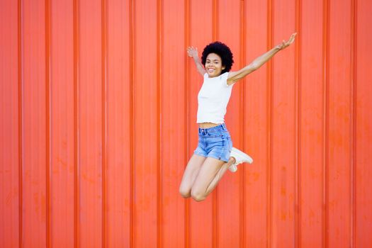 Full body of smiling African American female in casual outfit looking at camera while jumping with outstretched arms and bent knees against red background