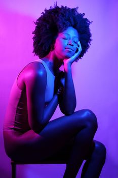 Side view of slim African American female model in bodysuit, with curly hair touching shoulder and looking away while sitting on stool under violet neon light