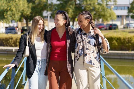 Group of content diverse female friends strolling on footbridge crossing river on sunny summer day in city with green trees