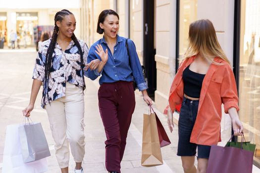 Positive multiracial female friends with shopping bags looking at each other and greeting woman while walking together on street near building after shopping in city