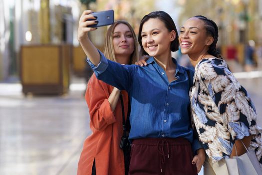 Group of cheerful diverse female friends taking self portrait on cellphone while standing on sidewalk against blurred background in city