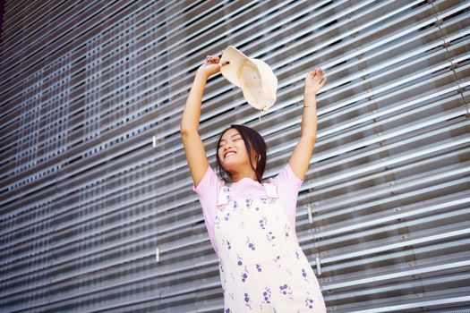 From below happy young Asian female in stylish clothes, smiling with closed eyes and raising arms with cap while dancing against metal wall on city street