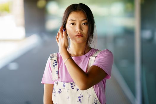 Young Asian woman in pink t shirt and stylish overalls, looking at camera and touching dark hair on blurred background of street and modern building on sunny day