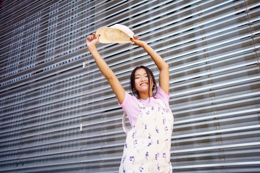 Low angle of optimistic young Asian woman in stylish clothes, raising arms with cap and looking at camera with smile against metal wall in daytime on city street
