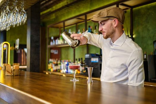 Bearded man in white shirt and cap filling glass with cocktail from shaker while standing behind counter and working in bar