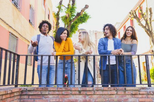 Company of multiethnic friendly people standing near railing in city and talking to each other while spending weekend together