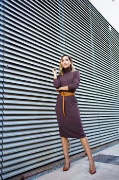 Full body self assured businesswoman in stylish dress with belt and high heeled shoes looking away while standing outside modern building on city street