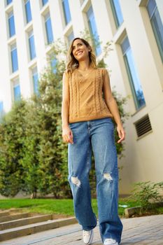 Low angle of glad woman in stylish clothes smiling and looking away while strolling on sidewalk outside modern building