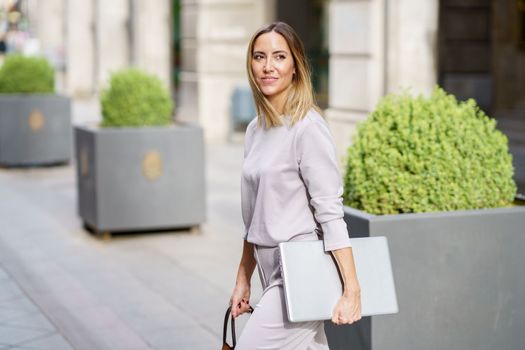 Side view of self assured adult female entrepreneur with blond hair in classy clothes, smiling and looking away while walking on city street with laptop in hand