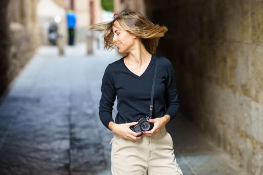 Positive young female traveler in casual clothes with vintage photo camera, smiling and shaking hair while standing on narrow path during sightseeing tour in old town