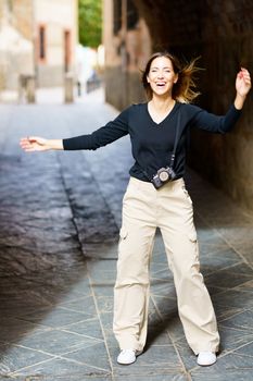 Full body of excited young female with long brown hair in casual clothes, smiling happily while standing in archway on old city street with photo camera during sightseeing trip