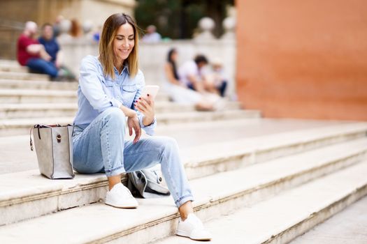Full body cheerful female in casual clothes smiling and browsing social media on cellphone while sitting on stairs near bag in daytime on city street