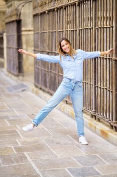 Full body cheerful adult female in casual clothes raising arm and leg and looking at camera with smile, while grasping bar of aged fence on city street