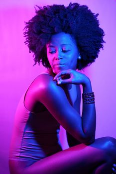 Side view of black woman in bodysuit with Afro hairstyle touching chin and looking away over shoulder while sitting under neon light against purple background