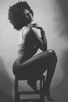 Side view of slim African American female model in bodysuit, with curly hair touching shoulder and looking away while sitting on stool. Black and white photograph.