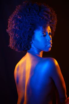 Sensual nude African American female with curly hair looking away over shoulder while standing under colorful neon illumination against black background