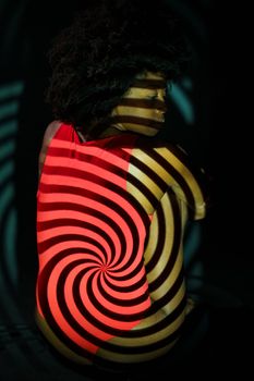 African American model in red bodysuit embracing knees and sitting under spiral projection in dark room