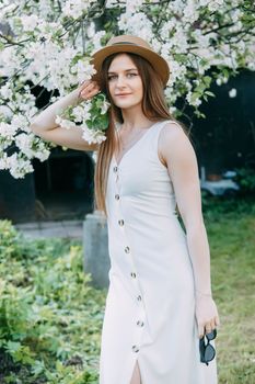 Beautiful young girl in white dress and hat in blooming Apple orchard. Blooming Apple trees with white flowers