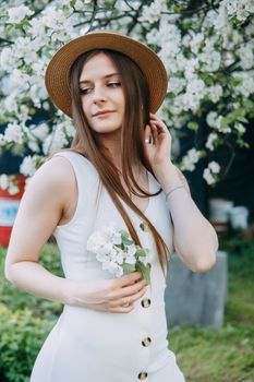 Beautiful young girl in white dress and hat in blooming Apple orchard. Blooming Apple trees with white flowers