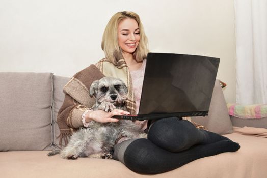 Lovely blond woman smiles and hugging a cute dog on the couch while watching movies on a laptop.