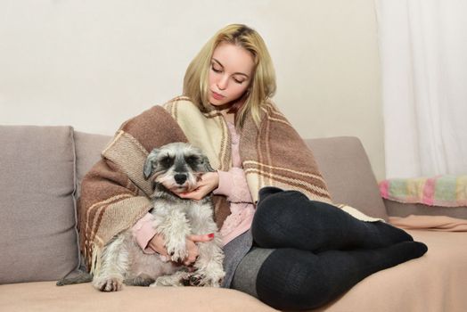 A beautiful blonde woman in a warm blanket sits comfortably on the sofa hugging her cute dog