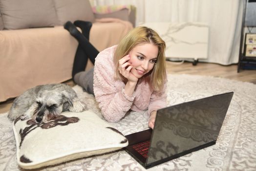 Lovely young lady in a pink sweater is lying on the carpet with a laptop at home, a gray dog is sleeping on a pillow next to her.
