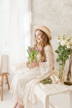 A girl with long hair in a dress and hat is sitting at the Easter table with cakes, spring flowers and quail eggs. Happy Easter celebration